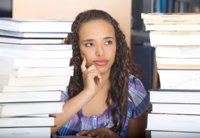 A woman with long hair looks thoughtful between two tall stacks of books.